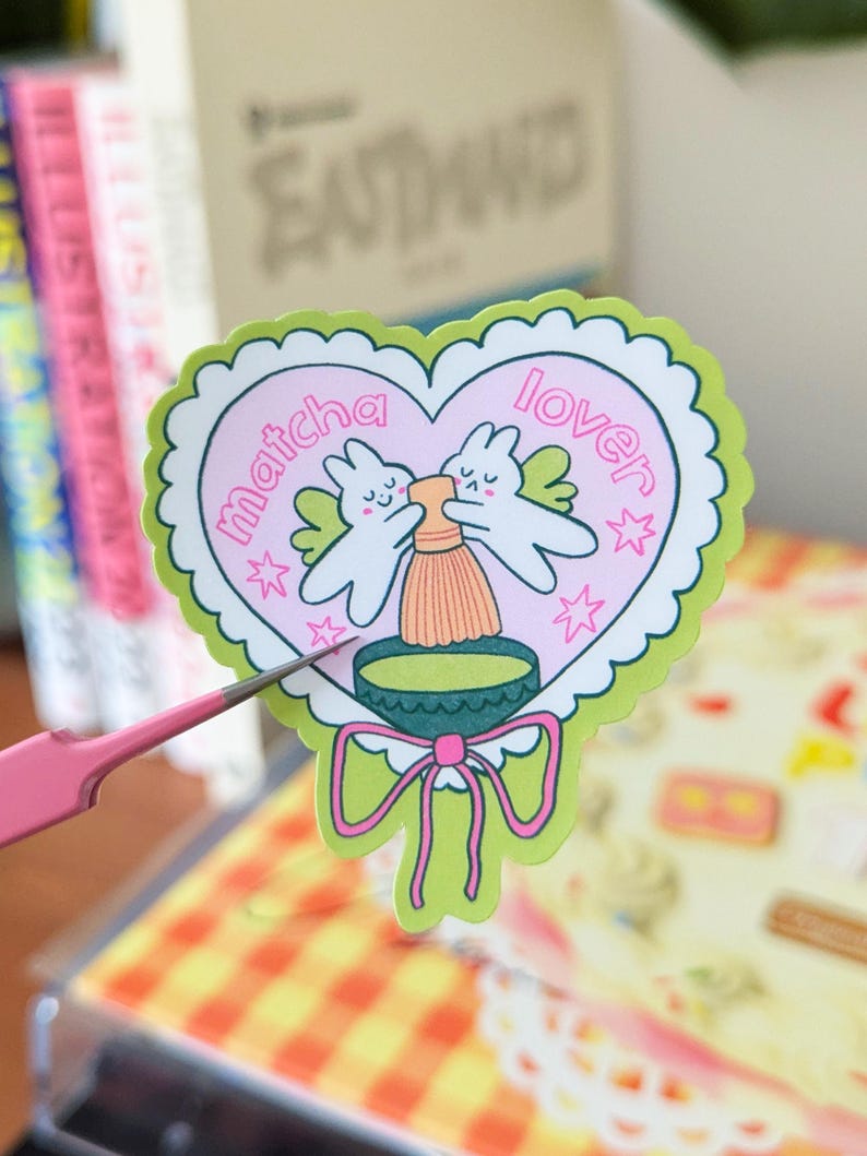 Puede incluir: Una pegatina en forma de coraz&oacute;n con un borde festoneado verde y un interior rosa. La pegatina presenta dos conejitos de dibujos animados con alas, un batidor de matcha y las palabras "matcha lover". Una cinta verde en la parte inferior.