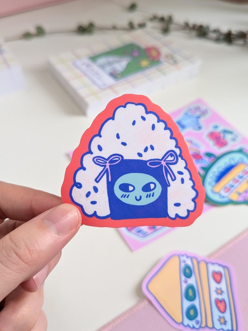 May include: A red, white, and blue cartoon sticker of a rice ball with a cute face. The rice ball has a blue and white patterned design and two pink bows.