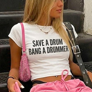 May include: White crop top with the text "SAVE A DRUM BANG A DRUMMER" in black. The top is paired with pink pants and a pink purse. A black shoulder bag is also visible.