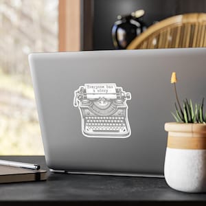 May include: A sticker featuring a vintage typewriter illustration in black and white, with the text "Everyone has a story." The sticker is affixed to a gray laptop, with a small potted plant in the foreground.