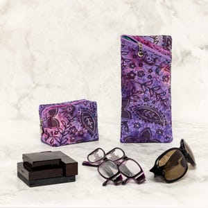 May include: Two purple and black patterned fabric eyeglass cases, one small and one large, with a pair of glasses and a black case on a white marble surface.