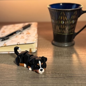 Może przedstawiać: Czarno-brązowa figurka bernardyna, śpiąca z głową opartą na łapach. Figurka wykonana jest z gładkiego, twardego materiału i ma realistyczny wygląd.