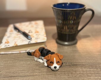 May include: A brown, black, and white dog figurine is lying on a wooden surface. A patterned notebook with a pen and a dark blue and black mug are in the background. The mug has a blue interior.