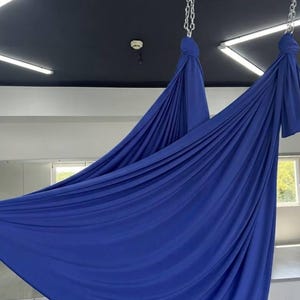 Könnte beinhalten: Eine tiefblaue Aerial-Yoga-Hängematte aus Stoff, die mit zwei silbernen Metallketten an der Decke befestigt ist. Der Stoff ist an den oberen Befestigungspunkten gerafft und hängt in einer großen, breiten und geschwungenen U-Form nach unten.