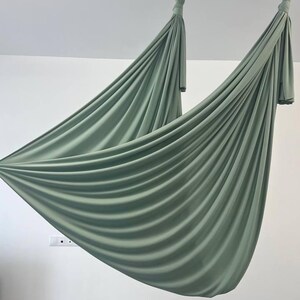 May include: Two sage green aerial yoga hammocks suspended from the ceiling. The fabric is draped and gathered, creating soft folds. The hammocks are attached to the ceiling with metal hardware, and hang in front of a white wall.