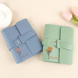 Könnte beinhalten: Zwei personalisierte Leder-Notizbücher mit Blumendesigns und den Namen "Nicola" und "Amrita" auf den Deckeln. Die Notizbücher sind blau und grün.