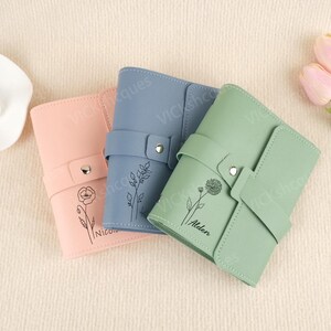 Könnte beinhalten: Drei personalisierte Leder-Notizbücher mit Blumendesigns und Namen. Die Notizbücher sind in Rosa, Blau und Grün.