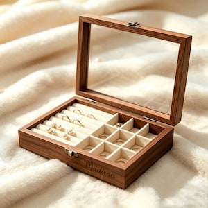 Könnte beinhalten: Eine rechteckige Schmuckschatulle aus Holz mit einem klaren Glasdeckel und einem Metallverschluss. Das Innere ist mit cremefarbenem Samt ausgekleidet und verfügt über Ringrollen sowie quadratische Fächer. Der Name Madison ist eingraviert.