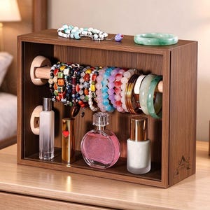 Könnte beinhalten: Ein rechteckiger Schmuck-Organizer aus Holz mit einer horizontalen Stange, an der verschiedene Perlenarmbänder und Armreifen hängen. Auf der unteren Ablage stehen ein durchsichtiger Parfümflakon, ein goldener Lippenstift mit rotem Herz und zwei Flaschen.