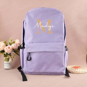 Könnte beinhalten: Ein lila Rucksack mit einer Vordertasche und schwarzen Trägern. Der Rucksack hat einen goldenen Buchstaben "M" mit dem Namen "Madge" in einer Schreibschrift. Der Hintergrund ist ein zartes Rosa.