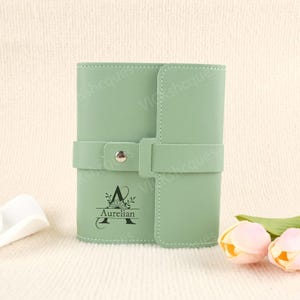 May include: A light green leather journal with a flap closure and a strap. The journal features the name "Aurelian" and the letter "A" with a floral design. The journal is on a white surface with two pink and yellow flowers.
