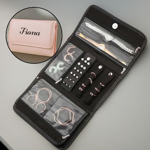 May include: Black jewellery roll with clear pockets and black velvet ring holders. The roll is open, displaying various earrings, rings, and a watch. A small pink jewellery case with the name "Fiona" is in the upper left corner.