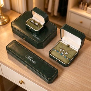 Könnte beinhalten: Ein Set aus dunkelgrünen Schmuckkästchen mit goldfarbenen Verzierungen und dem eingravierten Namen Maddie auf den Deckeln. Die Kollektion umfasst eine rechteckige Schatulle, eine lange schmale Box und eine offene Ringbox mit zwei Ringen und einer Kette.
