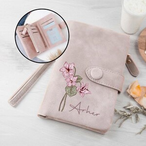 Könnte beinhalten: Eine rosa Ledergeldbörse mit einem Blumendesign und dem Namen "Arher" auf der Vorderseite geprägt. Die Geldbörse hat einen Druckknopfverschluss und ein Reißverschlussfach.