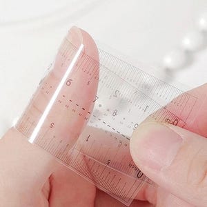 Pu&ograve; includere: Un righello trasparente e flessibile con marcature in pollici e centimetri. Il righello &egrave; tenuto in mano, mostrando la sua flessibilit&agrave;. I segni neri consentono misurazioni precise. Il righello misura circa 15 cm.