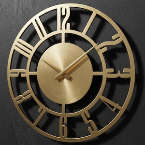 Pode incluir: Um relógio de parede dourado com algarismos romanos. O relógio tem um mostrador circular com acabamento em metal escovado e um design recortado. Os ponteiros são finos e dourados, indicando as horas. O relógio está montado numa parede cinza escuro.