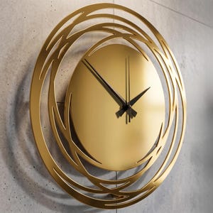 Peut inclure: Une horloge murale dorée avec un design circulaire. Le cadran de l'horloge est de couleur or uni avec des aiguilles noires. L'horloge est entourée d'un cadre décoratif ajouré. L'horloge est fixée sur un mur gris.