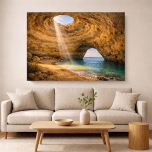 Op de afbeelding: Een rechthoekige wandkunstprint met een zonovergoten zeegrot met een natuurlijke opening in het plafond. Een lichtstraal schijnt op een zandstrand en turquoise water, met een grote rotsboog die uitkijkt op de horizon van de oceaan.