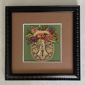 May include: A framed cross-stitch embroidery depicting a brown basket filled with a bouquet of autumn leaves in shades of orange, red, and yellow. The basket is surrounded by green leaves and the background is a light brown.