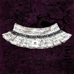 May include: A white and black musical note-printed skirt with a black lace trim and small, decorative embellishments. The skirt has a ruffled design and a small label with text.