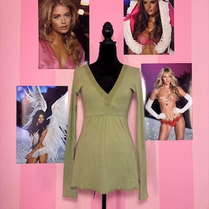 May include: A long-sleeved, olive green top with a V-neck and lace trim. The top is displayed on a black mannequin against a pink and white striped background. Photos of models are in the background.
