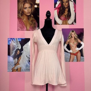 May include: A light pink long-sleeve top with a V-neckline and a decorative trim. The top is displayed on a black mannequin. The background features pink and white striped wallpaper and several photographs of models.