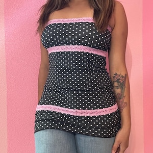 May include: A black tube top with white polka dots and pink lace trim. The top is sleeveless and form-fitting, with a straight neckline. The pink lace trim is at the top and mid-section of the top. The top is paired with blue jeans.