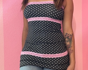 May include: A black tube top with white polka dots and pink lace trim. The top is sleeveless and form-fitting, with a straight neckline. The pink lace trim is at the top and mid-section of the top. The top is paired with blue jeans.