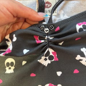 以下が含まれることがあります： ピンクのリボンとハートが付いた白いドクロ模様の黒いトップス。トップスは細いストラップと装飾ボタンが付いています。背景のタグには赤い唇のプリントと「Kiss's」の文字があります。