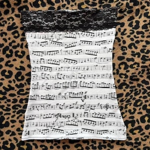 Puede incluir: Un top de tubo blanco con un ribete de encaje negro en el borde superior. La parte superior está cubierta con una impresión de partitura musical negra. El fondo es un estampado de leopardo marrón y beige.