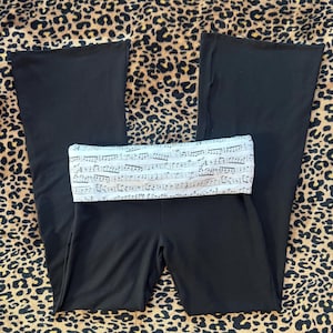 Puede incluir: Pantalones de yoga negros con una cintura ancha y plegable. La cintura presenta una tela blanca estampada con notas musicales y pentagramas. Los pantalones tienen un diseño de pierna acampanada, adecuado para danza o yoga.