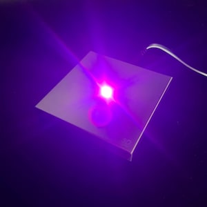 May include: A square, metallic object emitting a bright, purple light. The light source is centered, creating a starburst effect. A thin, white cable extends from the object. The object has the text "3D" on the corner.