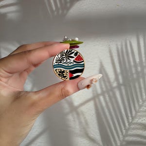 May include: A round enamel pin with a colourful design featuring a watermelon, palm trees, and a beach scene. The pin is gold-toned and has a white background.