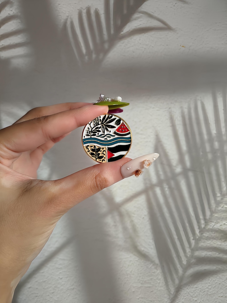 May include: A round enamel pin with a colourful design featuring a watermelon, palm trees, and a beach scene. The pin is gold-toned and has a white background.