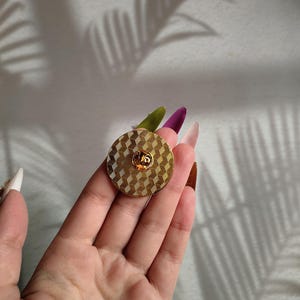 May include: A round pin with a geometric pattern, finished in gold. The pin has a textured surface and a small, raised design in the centre.