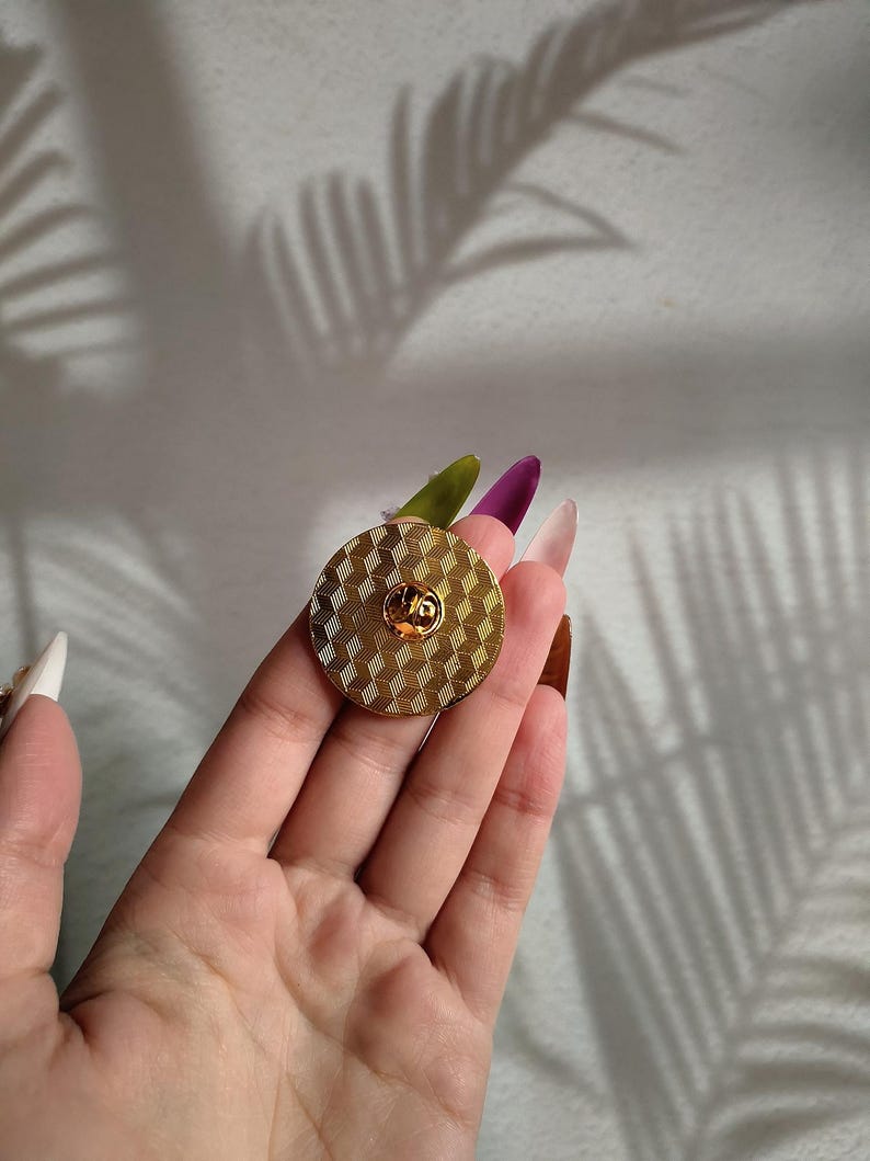 May include: A round pin with a geometric pattern, finished in gold. The pin has a textured surface and a small, raised design in the centre.