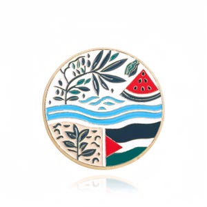 May include: A round, gold-coloured pin with a white background and colourful designs. The pin features a stylised image of a tree, a river, a watermelon, and a flag. The flag is red, black, and green with a white triangle in the top left corner.