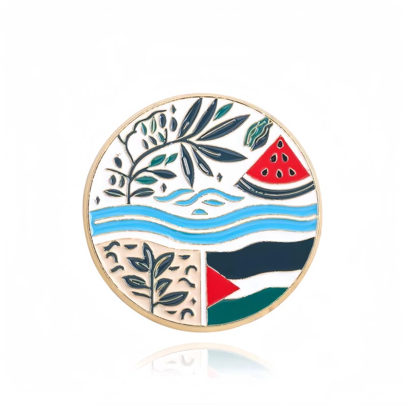 May include: A round, gold-coloured pin with a white background and colourful designs. The pin features a stylised image of a tree, a river, a watermelon, and a flag. The flag is red, black, and green with a white triangle in the top left corner.
