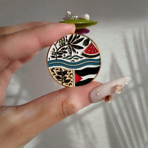 May include: A round enamel pin with a white background and a colourful design. The design features a watermelon slice, a palm tree, and a blue and white striped wave. The pin has a gold metal finish.