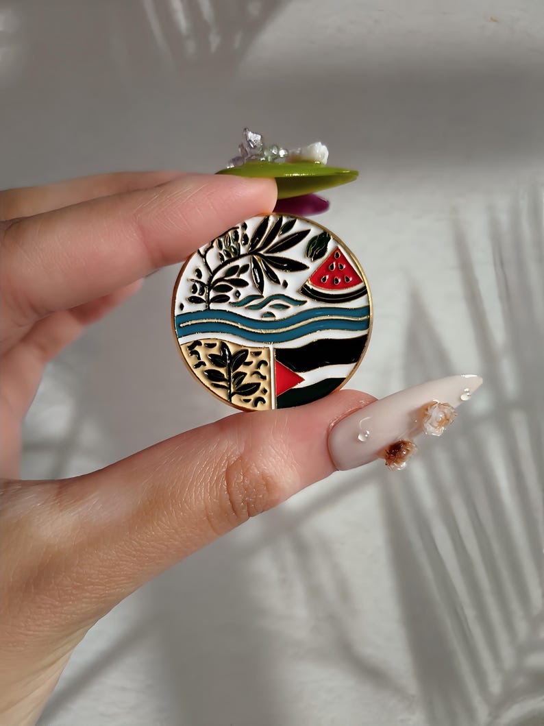 May include: A round enamel pin with a white background and a colourful design. The design features a watermelon slice, a palm tree, and a blue and white striped wave. The pin has a gold metal finish.