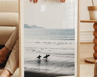 May include: A framed photograph of two people walking on a beach with surfboards. The beach is sandy and the water is blue. The sky is cloudy and the horizon is in the distance.
