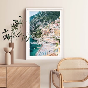 May include: A colorful photograph of a town built on a cliffside overlooking a beach with blue water. The town is a mix of yellow, orange, and red buildings. The beach is crowded with people and umbrellas.