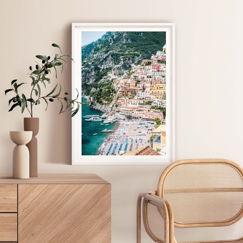 May include: A colorful photograph of a town built on a cliffside overlooking a beach with blue water. The town is a mix of yellow, orange, and red buildings. The beach is crowded with people and umbrellas.
