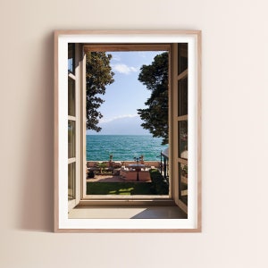 May include: A view of a lake through an open window with a wooden frame. The lake is blue and there are mountains in the distance. There are trees on either side of the window and a patio with tables and chairs in the foreground.