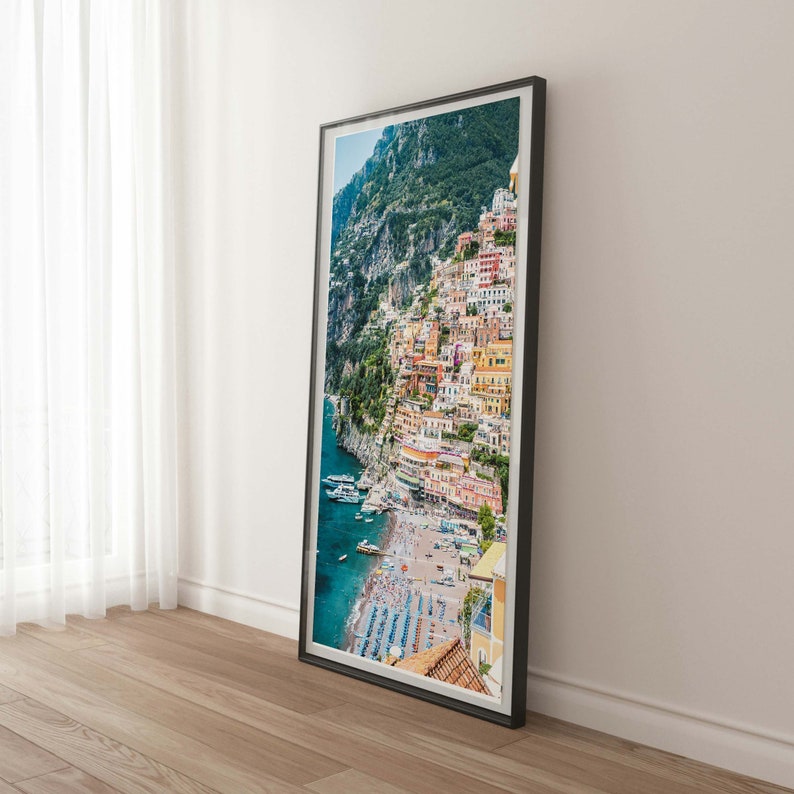 May include: A framed photograph of a colorful Italian village built into a cliffside overlooking a blue ocean. The village is filled with buildings in shades of yellow, orange, red, and pink. The ocean is dotted with boats and the beach is lined with umbrellas.