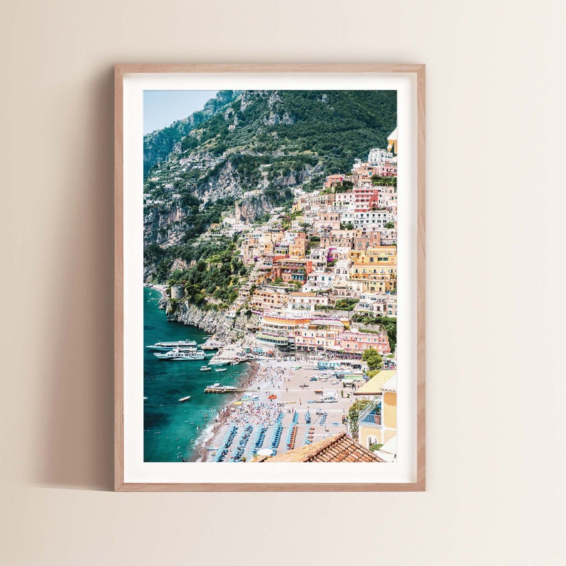 May include: A colorful cityscape on a cliffside overlooking a beach with turquoise water and boats. The buildings are a mix of yellow, pink, orange, and white, with green trees and foliage.