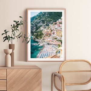 May include: A colorful photograph of a coastal town built into a hillside overlooking a beach with turquoise water. The town is a mix of yellow, orange, pink, and red buildings. There are boats in the water and people on the beach.