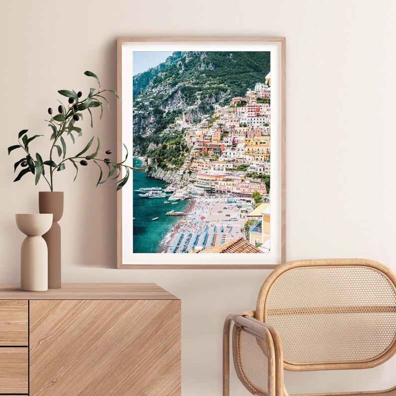 May include: A colorful photograph of a coastal town built into a hillside overlooking a beach with turquoise water. The town is a mix of yellow, orange, pink, and red buildings. There are boats in the water and people on the beach.