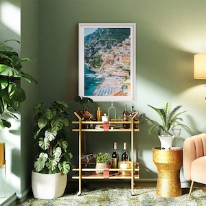 May include: A gold bar cart with two shelves, a white planter with a green plant, a gold side table with a white planter and a green plant, and a framed print of a coastal town with colorful buildings.
