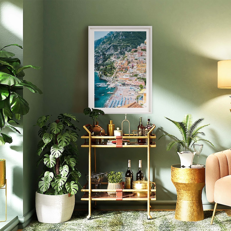 May include: A gold bar cart with two shelves, a white planter with a green plant, a gold side table with a white planter and a green plant, and a framed print of a coastal town with colorful buildings.